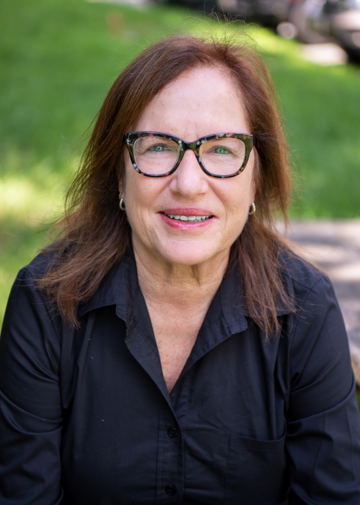 A headshot of a woman with shoulder length brown hair and glasses. She is wearing a black button up and smiling at the camera.